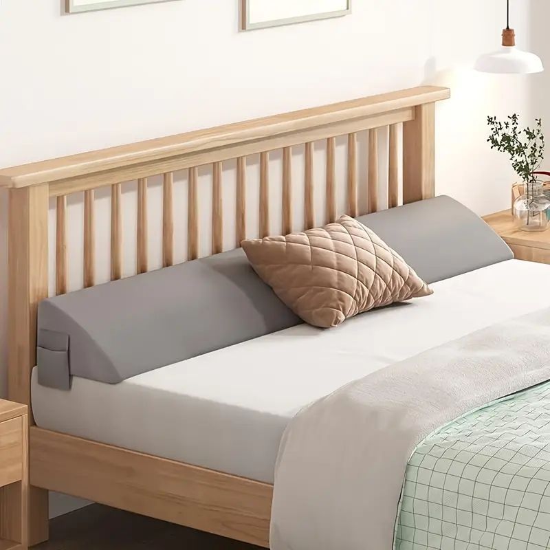 Product photograph of a long gray triangular cushion between a mattress and wooden headboard
